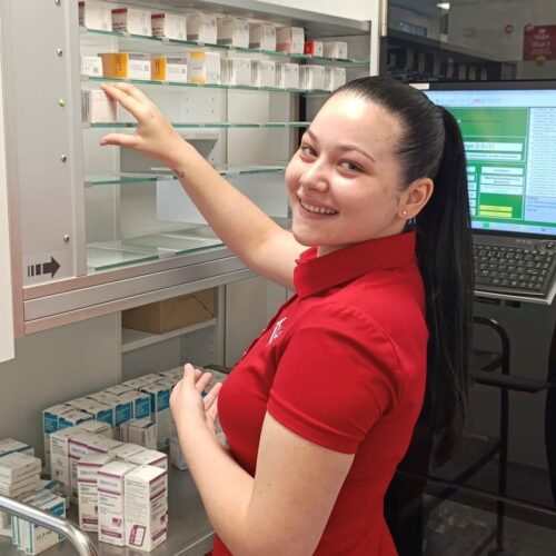 Mitarbeiterin in rotem Apotheken-Poloshirt sortiert Medikamente in einem Apothekenschrank und lächelt in die Kamera.